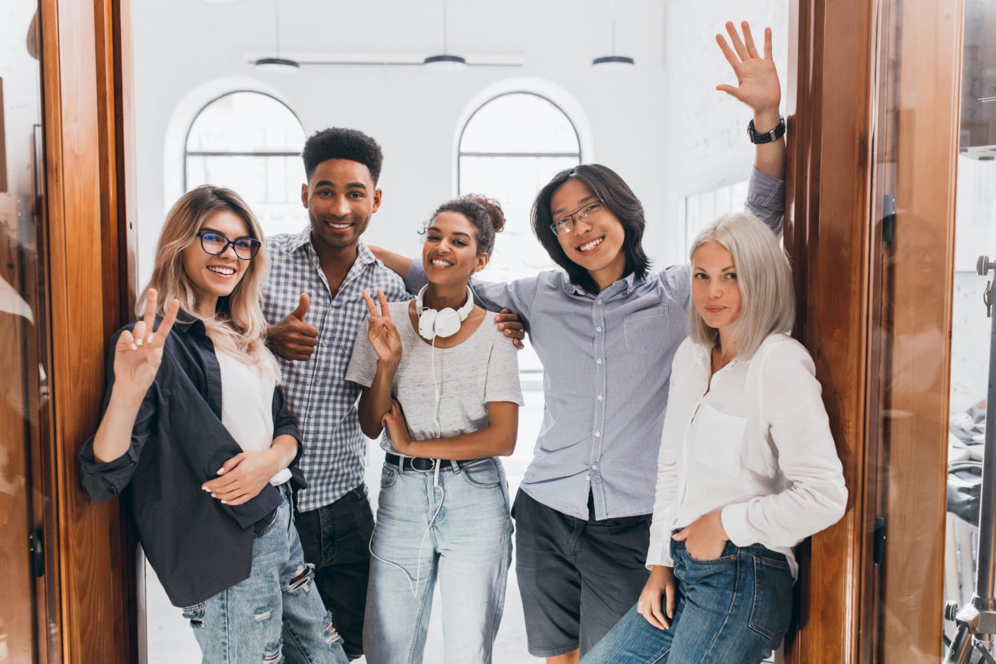 Slim african girl in jeans and white headphones standing in office with colleagues. European freelance female programmer having fun with friends and posing with pleasure.